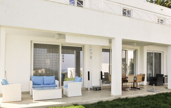 A modern building with a covered outdoor seating area featuring white sofas and chairs, a dining table, and a small table with a candle.
