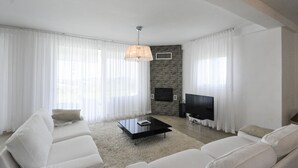 Ein modernes Wohnzimmer mit einem großen Flachbildfernseher, weißen Sofas, einem Couchtisch und transparenten Vorhängen.