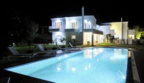 A modern house with a swimming pool, lounge chairs, and a well-lit exterior at night.