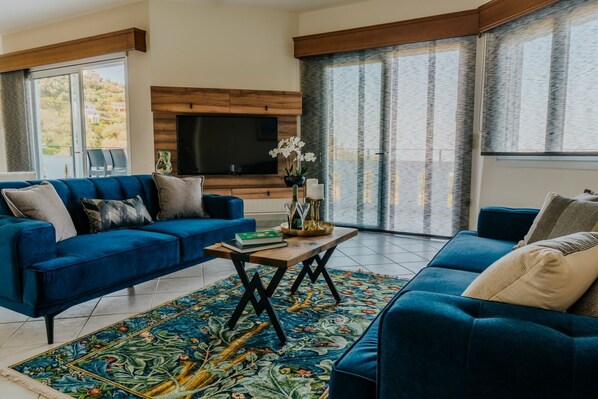 A modern living room with a blue sofa set, a flat-screen TV mounted on a wooden wall, and a large window with curtains. - Villa Agapios - Six Bedroom Villa, Sleeps 11 (Agios Dimitrios Rodias)