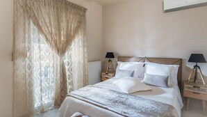 Un dormitorio con una cama grande, dos mesitas de noche con lámparas, un radiador y una ventana con cortinas traslúcidas.