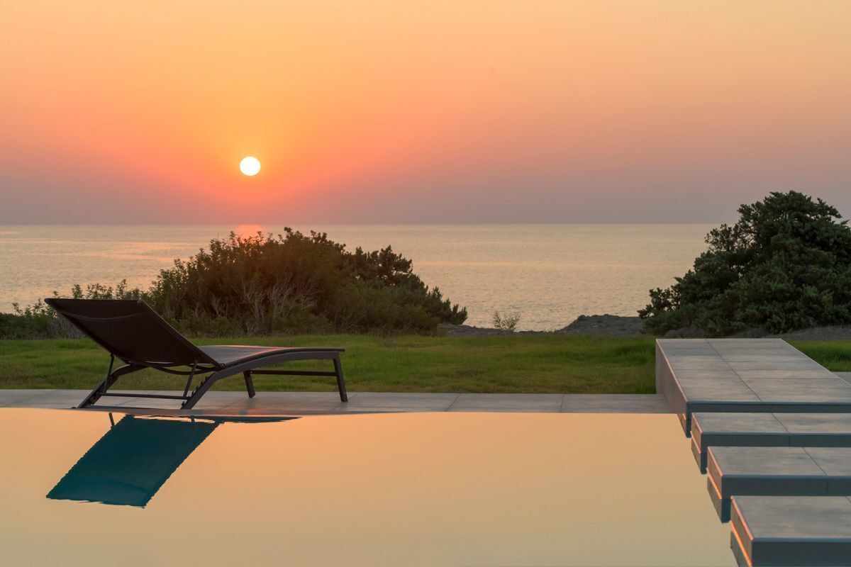 An infinity pool with a lounge chair and a bench, overlooking a sunset over the sea.