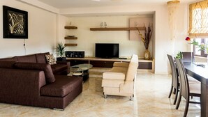 Un salon moderne avec un canapé marron, un fauteuil beige, une table basse en verre et un téléviseur à écran plat. On y trouve également des étagères décoratives et un lustre.