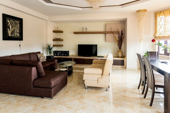 A modern living room with a brown sofa, a beige armchair, a glass coffee table, and a flat-screen TV. There are decorative shelves and a chandelier.
