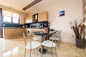 A modern kitchen with a dining area, featuring a round glass table with four chairs, a built-in oven, and a large window.