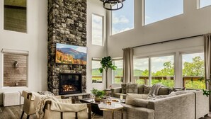 A spacious living room with a stone fireplace, comfortable seating, and large windows offering a view of the outdoors. - Swim Spa/Views/Gameroom/Theater/Luxe Design - Rabbit Run (Gatlinburg)