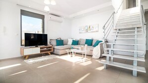 Een moderne woonkamer met een bank, salontafel en tv-meubel. Een trap met een leuning en een groot raam zorgen voor veel daglicht.
