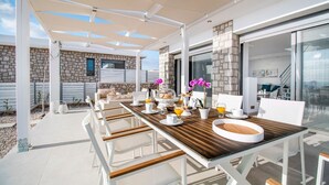 Een overdekt terras met een houten eettafel gedekt voor het ontbijt, witte stoelen en een stenen gebouw op de achtergrond.