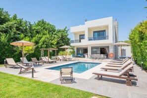 Une maison moderne à deux étages, dotée d’une piscine, de chaises longues et de parasols.