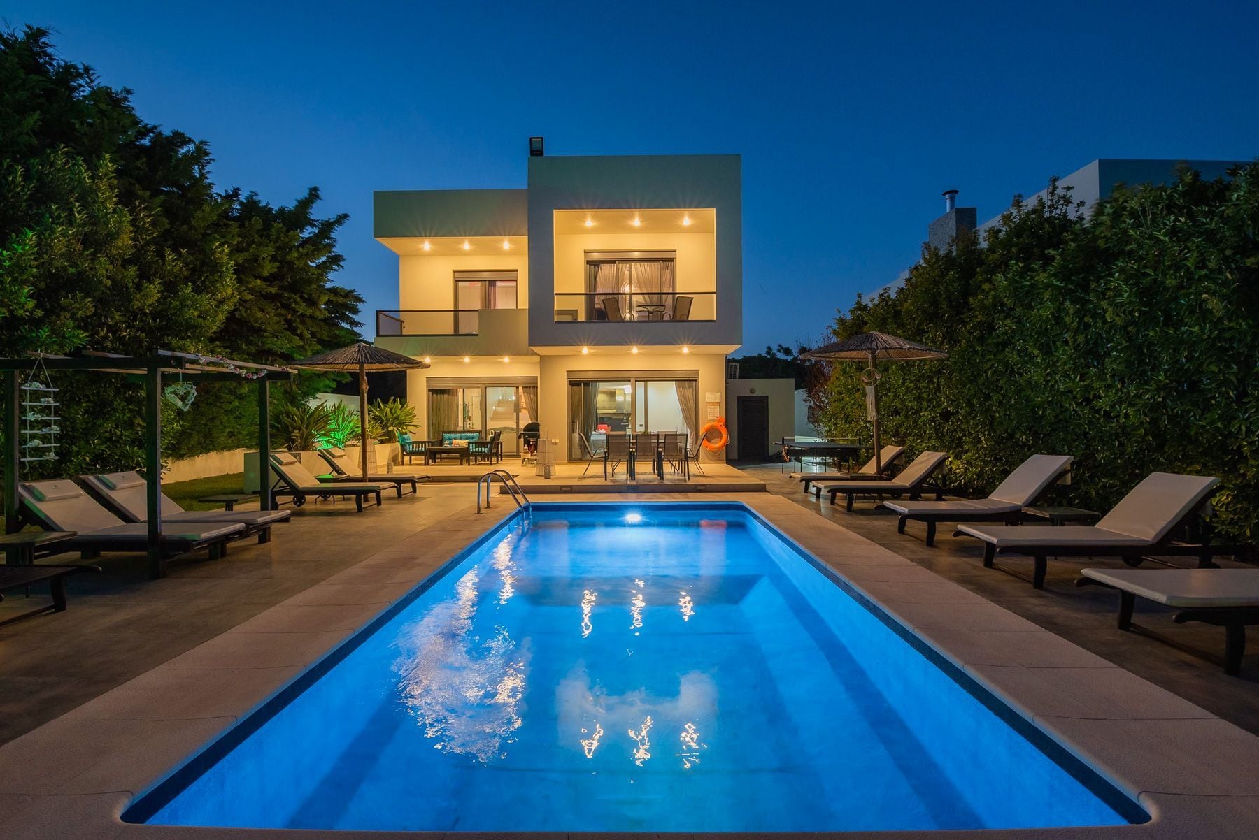A modern two-story house with a swimming pool, outdoor seating, and a well-lit patio area.