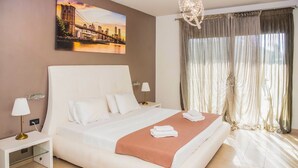 Una camera d'albergo con un letto grande, un comodino, una lampada, un quadro che raffigura un paesaggio urbano e un'ampia finestra con tende.
