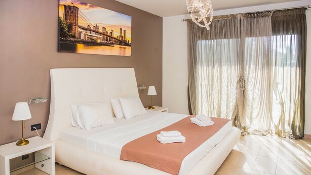 Une chambre d’hôtel avec un grand lit, une table de chevet, une lampe, un tableau représentant un paysage urbain et une grande fenêtre avec des rideaux.