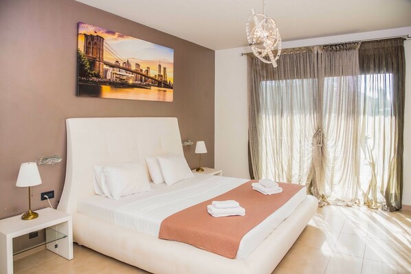 Une chambre d’hôtel avec un grand lit, une table de chevet, une lampe, un tableau représentant un paysage urbain et une grande fenêtre avec des rideaux.