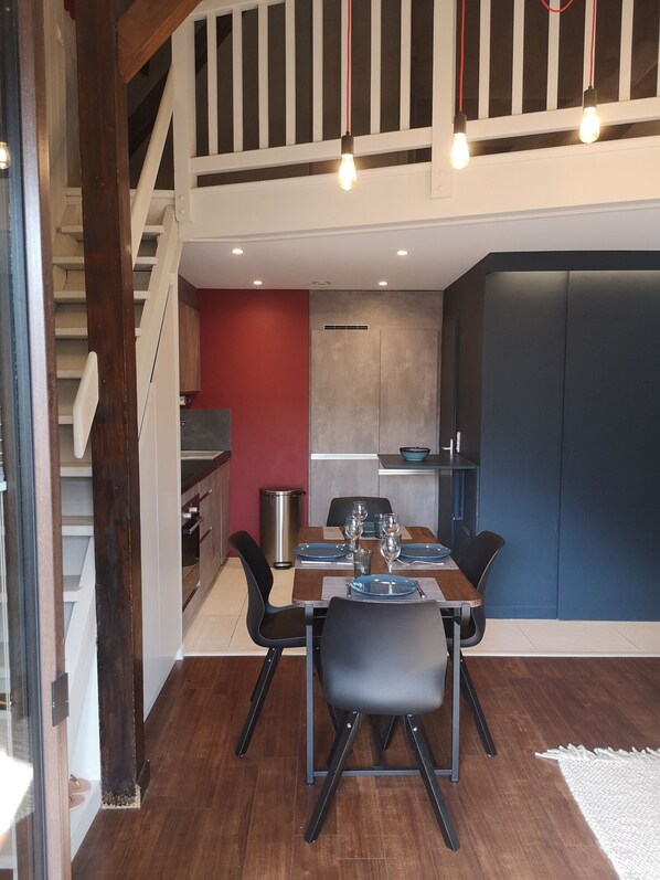 A dining area with a wooden table set for four, a red accent wall, and a view of a kitchen and staircase.