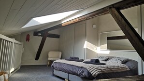 Un dormitorio con techo inclinado, una cama con varias mantas, una silla y un ventanal.