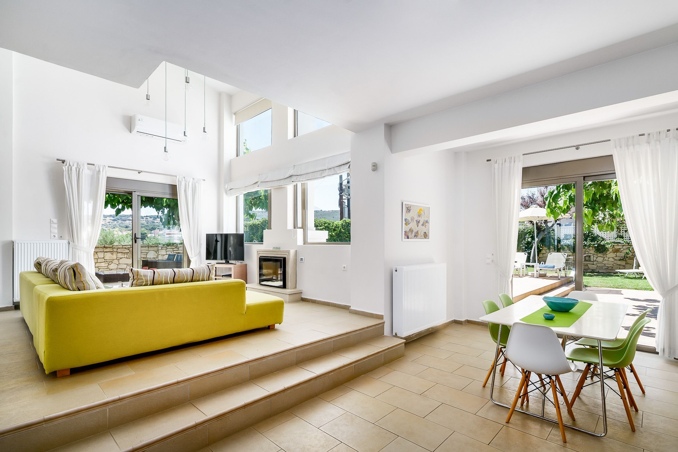 A bright living room with a yellow sofa, a TV, and a dining area with a table and chairs.