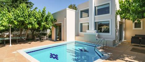 Ein moderner Poolbereich mit einem klaren blauen Pool, umgeben von einer Terrasse mit Gartenmöbeln und einem Gebäude mit großen Fenstern.