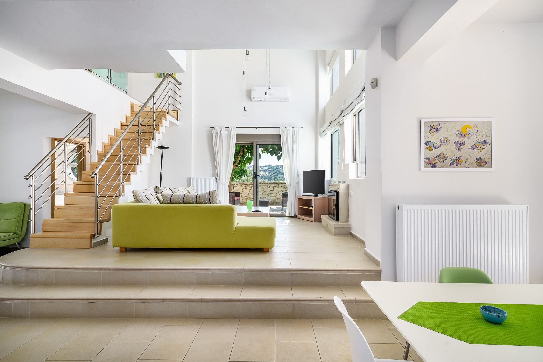 A modern living room with a staircase, a green sofa, and a dining area with a table and chairs.