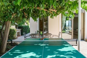Ein Tischtennis-Tisch im Freien unter einem Baum, umgeben von Stühlen und einem Tisch, mit einem Gebäude im Hintergrund.