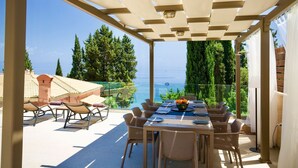 A covered outdoor dining area with a table set for four, overlooking a scenic view of the sea and greenery. - Empress Sissi - Seven Bedroom Villa, Sleeps 14 (Gastouri)
