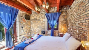 Ein Schlafzimmer mit einer Steinwand, einem Bett mit weißen Leinen, blauen Vorhängen und einer Holzbalkendecke.