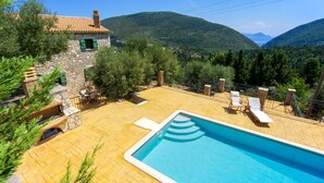 A swimming pool with lounge chairs, a stone building with a tiled roof, and a mountainous landscape in the background.