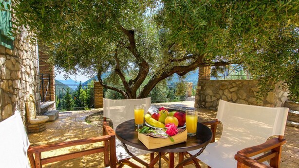A table with a fruit basket and drinks, surrounded by chairs, in a sunny outdoor setting with stone walls and a view of the mountains.