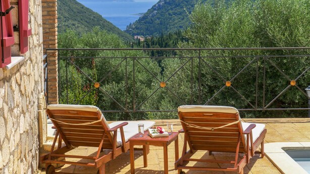 Un balcón con dos sillas de madera y una mesita pequeña, con vistas a un paisaje montañoso y una masa de agua.