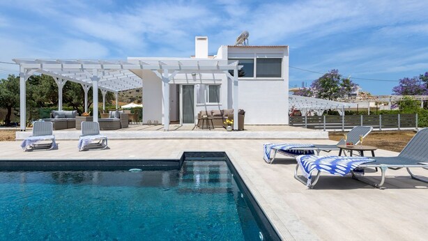 Ett modernt utomhusområde med en pool, solstolar, en pergola och en liten byggnad med vit fasad.