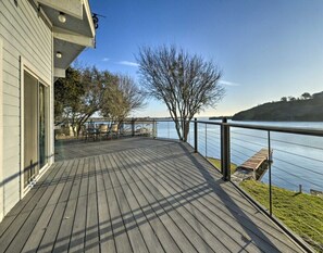 Une terrasse en bois avec une balustrade donnant sur une étendue d’eau, un ciel dégagé et un arbre en arrière-plan.