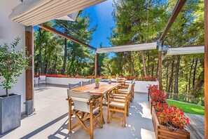 Eine Terrasse im Freien mit einem Holztisch und Stühlen, umgeben von Blumentöpfen und Blick auf Bäume.