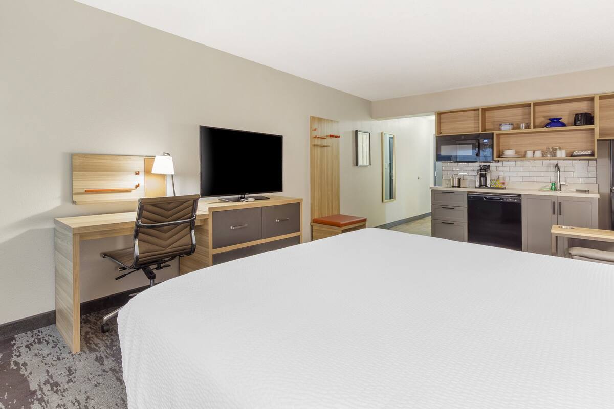 Ein Hotelzimmer mit einem großen Bett, einem Schreibtisch mit Stuhl, einem Fernseher, einer kleinen Küchenzeile und einem Badezimmer.