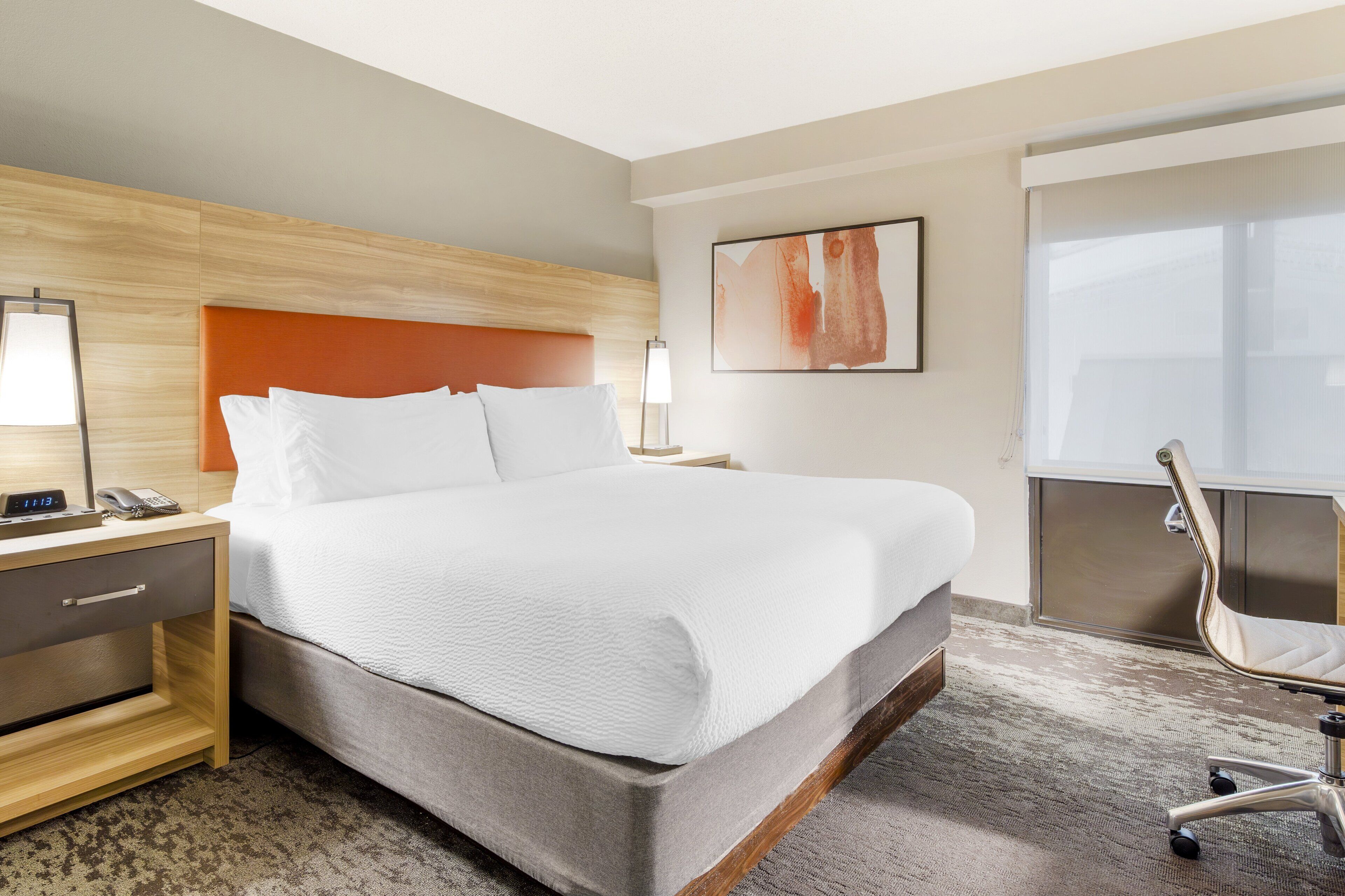 Une chambre d’hôtel avec un grand lit, une table de chevet avec une lampe, une chaise et une fenêtre avec des stores.