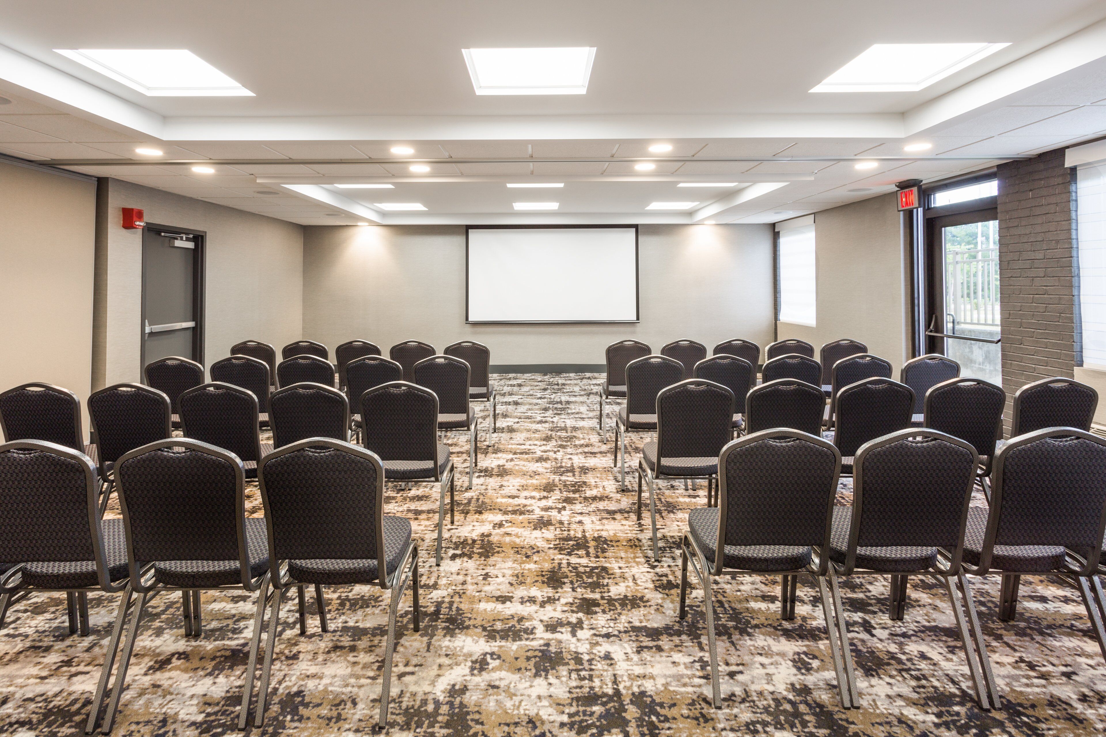 A conference room with rows of chairs facing a blank screen, carpeted floor, and a door with a red emergency exit sign.