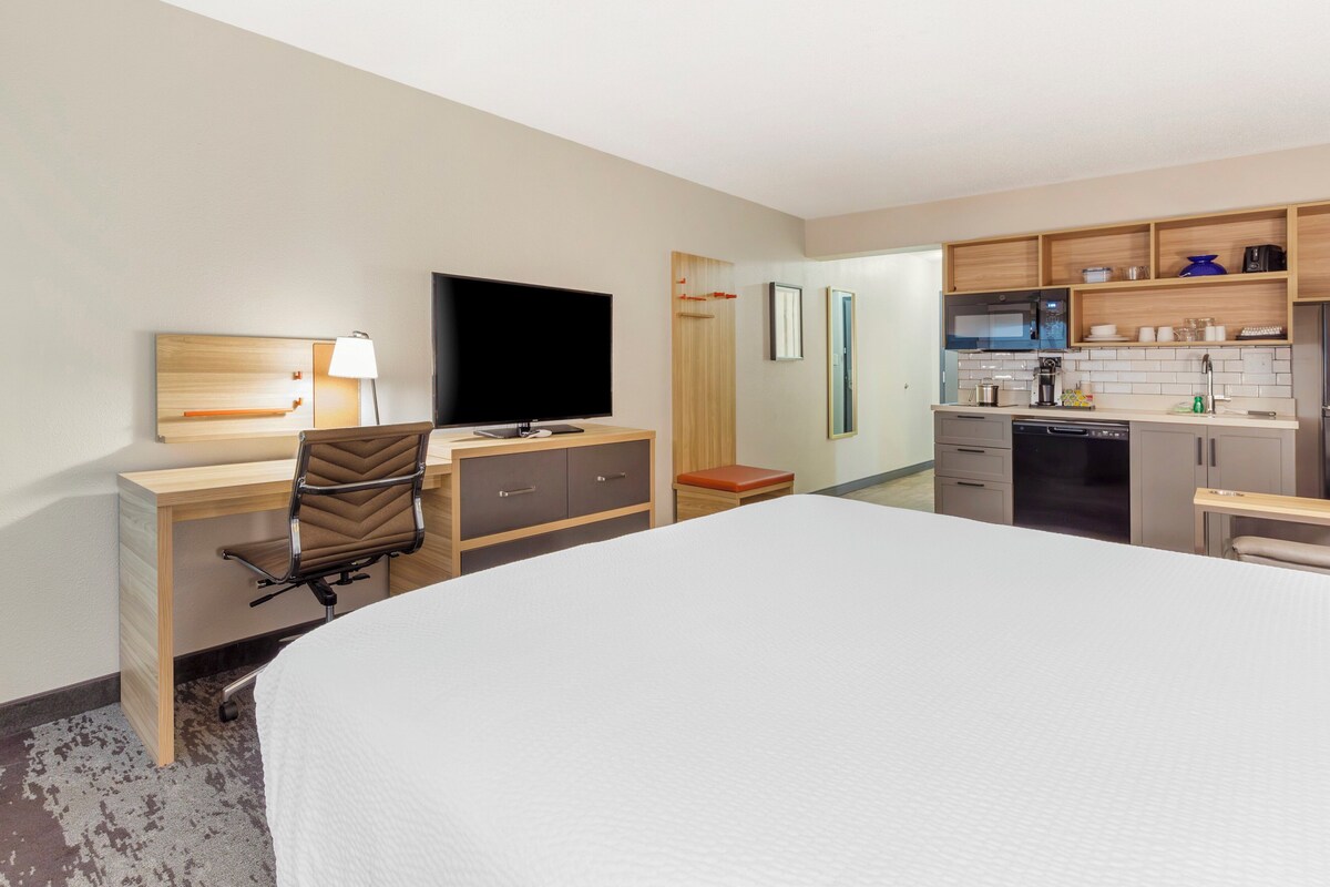 Quarto de hotel com uma cama grande, uma escrivaninha com cadeira, televisão, pequeno espaço de cozinha e banheiro.