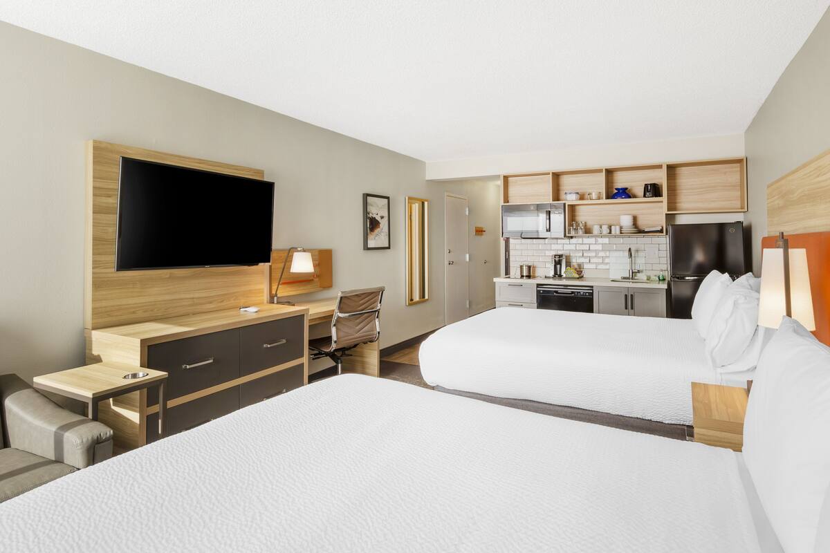 Ein Hotelzimmer mit zwei Betten, einem Schreibtisch, einem Stuhl, einem Fernseher und einer Küchenzeile.