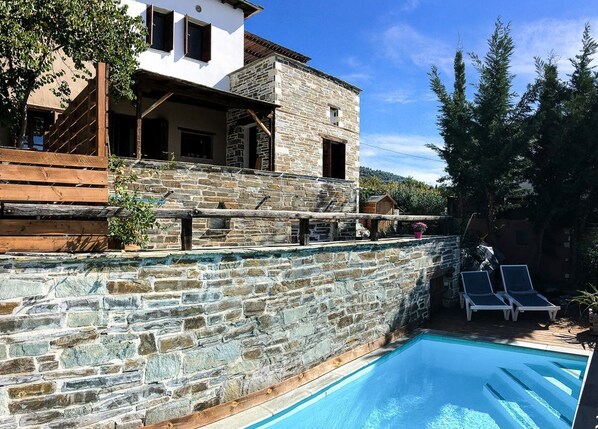 A stone-walled pool area with a clear blue pool, wooden deck, and lounge chairs. - Villa Artemida - Three Bedroom Villa, Sleeps 8 (Agios Georgios Nilias)