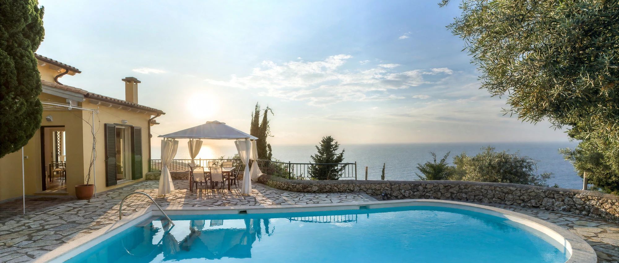 A swimming pool with a stone deck, a covered seating area, and a view of the sea.