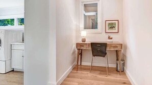 Una habitación con un escritorio de madera, una silla, una mesita de noche con una lámpara y un cuadro en la pared.