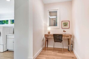 Una habitación con un escritorio de madera, una silla, una mesita de noche con una lámpara y un cuadro en la pared.