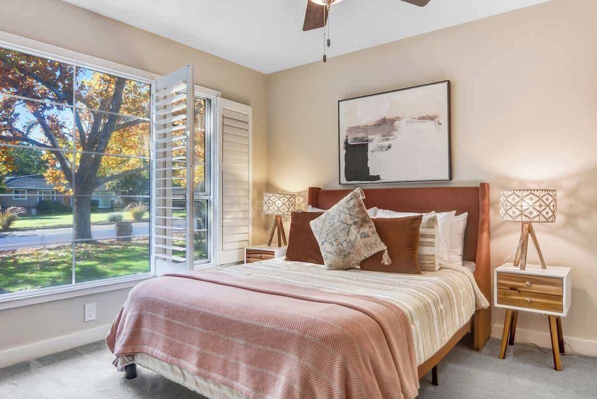 Um quarto com cama, janela com persianas, ventilador de teto, mesa de cabeceira com abajur e um quadro na parede.