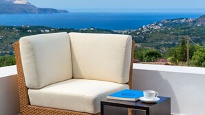 A wicker chair with a cushion, a book, and a cup on a small table, overlooking a coastal landscape with houses and cliffs.