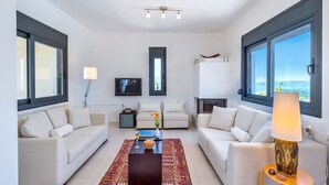 A modern living room with two white sofas, a coffee table, a TV, and large windows offering a view of the sea.