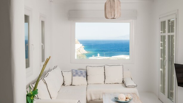 A white room with a view of the sea, a white sofa, and a hanging light fixture.