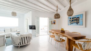 A bright, spacious room with a wooden dining table, white chairs, a white sofa, and a large window.