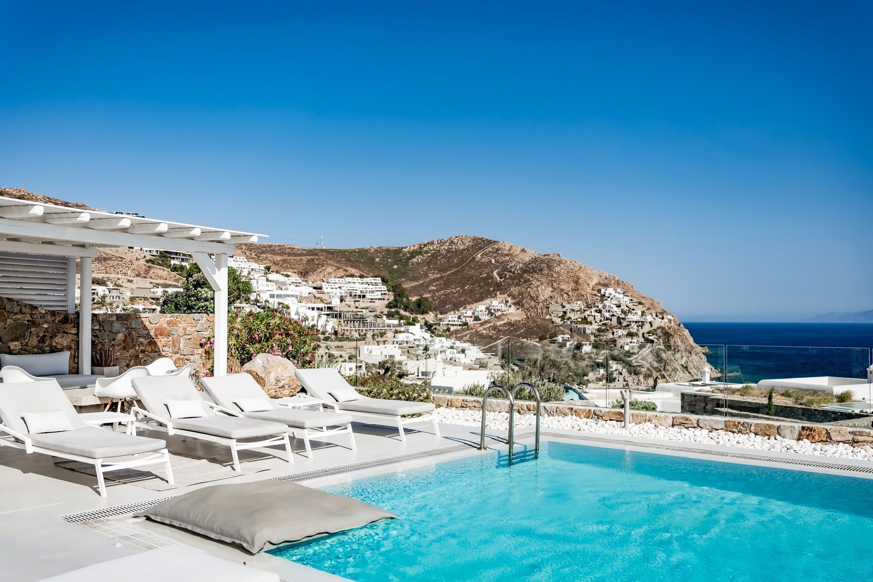 A poolside area with white lounge chairs and a clear view of a coastal town and the sea.