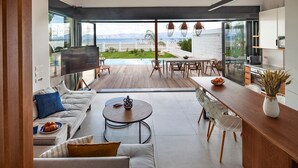 A modern living room with a large TV, a wooden dining table, and a view of a pool and mountains. - Villa Ammos - Two Bedroom Villa, Sleeps 6 (Kavos)