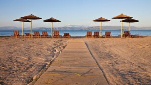 A beach with wooden lounge chairs and thatched umbrellas, a wooden pathway leading to the water, and a clear view of the sea and distant mountains. - Villa Ammos - Two Bedroom Villa, Sleeps 6 (Kavos)