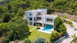 Une maison moderne à deux étages, dotée d’une piscine, entourée d’une végétation luxuriante et de collines.