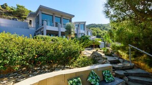 A modern two-story house with a balcony, surrounded by lush greenery and a stone retaining wall. - Villa Arthur - Four Bedroom Villa, Sleeps 8 (Rhodes)